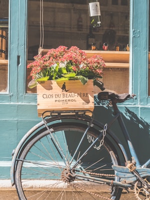 A vintage blue bicycle is parked against a teal-colored wall with a wooden crate attached to the back. The crate is filled with red flowers and bears the label 'Clos du Beau-Père Pomerol 2014'. In the background, a window displays bottles of wine, contributing to a rustic and charming ambiance.