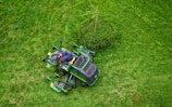 A person in a dark jacket and hat operates a green lawn mower on a lush, green grass field. The mower is maneuvering around a small bush, creating visible patterns on the lawn.
