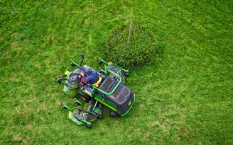 A person in a dark jacket and hat operates a green lawn mower on a lush, green grass field. The mower is maneuvering around a small bush, creating visible patterns on the lawn.