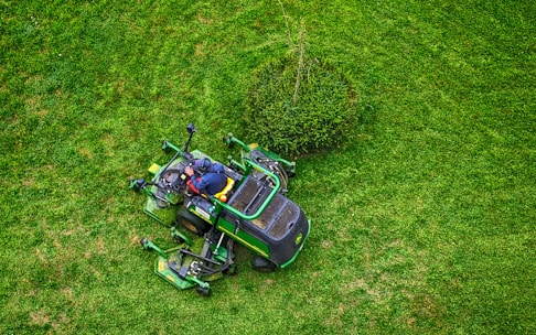 A person in a dark jacket and hat operates a green lawn mower on a lush, green grass field. The mower is maneuvering around a small bush, creating visible patterns on the lawn.