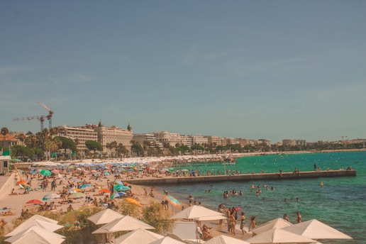 A crowded beach scene with numerous people relaxing under colorful umbrellas on the sand and swimming in the turquoise sea. In the background, a line of hotels and buildings is visible, with cranes indicating construction activity. Palm trees add to the tropical atmosphere under a clear blue sky.