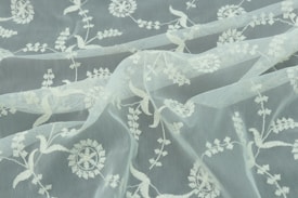 A delicate piece of fabric featuring intricate white floral embroidery on a sheer, translucent background. The fabric has gentle folds, showcasing the detailed patterns and the lightweight nature of the material.