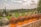 Large glass carboys filled with amber liquid are lined up on a stone ledge. In the background, there is a lush green vineyard with rows of grapevines stretching into the distance. The landscape is surrounded by tall trees and rolling hills under a partly cloudy sky.