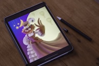 A digital artwork is displayed on a tablet placed on a wooden surface, with a stylus lying beside it. The artwork features a whimsical character wearing a hat and goggles, with a bushy beard and outstretched arm. Surrounding the character are stars, a crescent moon, and several hot air balloons, all set against a gradient background transitioning from blue to yellow.