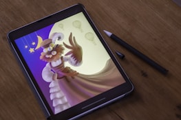 A digital artwork is displayed on a tablet placed on a wooden surface, with a stylus lying beside it. The artwork features a whimsical character wearing a hat and goggles, with a bushy beard and outstretched arm. Surrounding the character are stars, a crescent moon, and several hot air balloons, all set against a gradient background transitioning from blue to yellow.