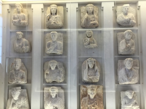 A collection of stone relief sculptures displayed within a glass case. Each relief depicts a human figure, mostly in regal or attentive postures. The sculptures appear to be ancient, with detailed carvings emphasizing clothing folds and facial features.