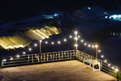 A wooden deck is illuminated by strings of warm fairy lights, creating a cozy and inviting atmosphere. In the background, greenhouses with glowing roofs add a serene ambiance. A small white table is present on the deck.