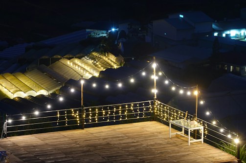 A wooden deck is illuminated by strings of warm fairy lights, creating a cozy and inviting atmosphere. In the background, greenhouses with glowing roofs add a serene ambiance. A small white table is present on the deck.
