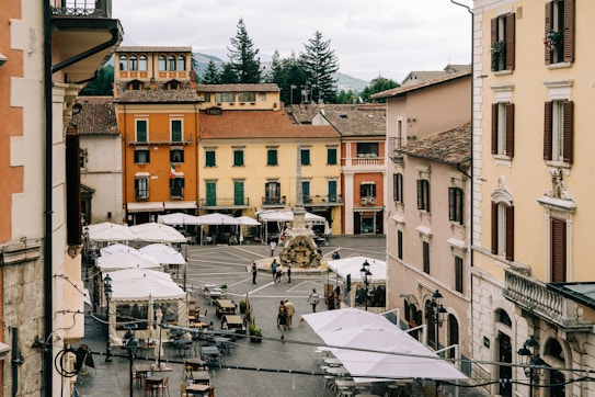A quaint town square framed by historic buildings with colorful facades and shuttered windows. The cobblestone plaza hosts several tables and umbrellas, suggesting a caf&eacute; or dining area. People are walking and gathering around a central fountain, adding life and activity to the scene.