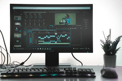 A computer monitor showing a video editing software interface, with timelines, video clips, and audio mixers visible. A keyboard and mouse are placed in front of the monitor on a clean desk. A small potted plant is situated on the right side, adding greenery to the workspace.