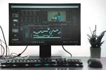 A computer monitor showing a video editing software interface, with timelines, video clips, and audio mixers visible. A keyboard and mouse are placed in front of the monitor on a clean desk. A small potted plant is situated on the right side, adding greenery to the workspace.
