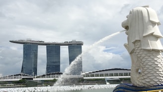 A large, iconic statue of a lion with a fish body spouts water into a bay. In the background, three tall towers support a boat-shaped structure on top, set against a cloudy sky.