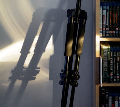 A tripod is leaning against a wall, casting a shadow that stretches across the surface. To the right, there is a bookshelf filled with DVDs or Blu-rays, with the titles clearly visible on the spines. The lighting creates a mix of highlights and shadows on the wall and the tripod.