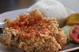 A plate of food featuring crispy fried chicken topped with a spicy red sauce, served alongside white rice. Slices of cucumber and a tomato garnish accompany the dish, along with a piece of fried yellow tofu.