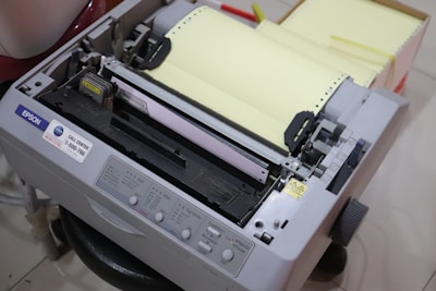 An Epson dot matrix printer with control buttons and a stack of continuous form paper loaded. The printer appears to be in an office or workspace setting.