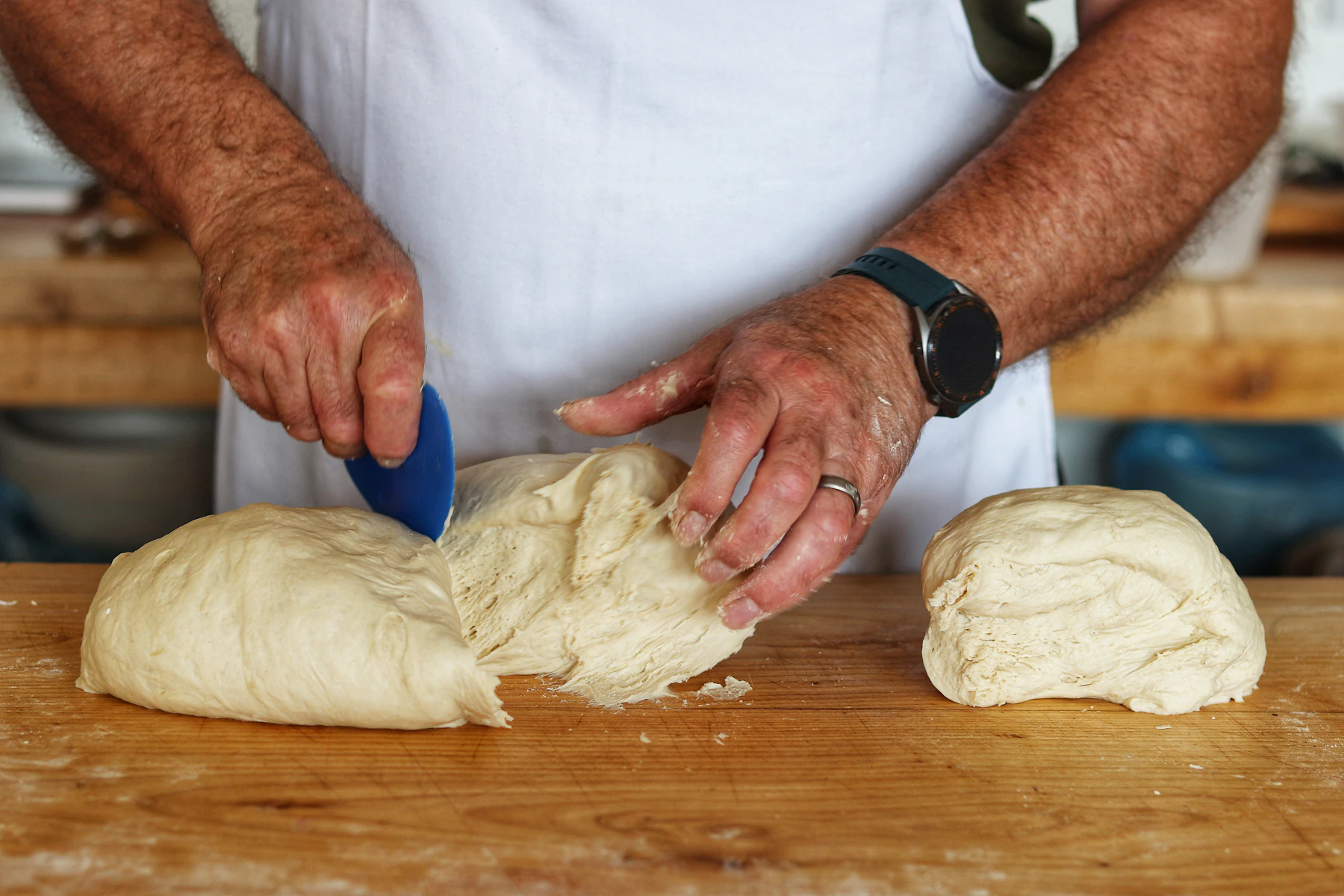 Hands dividing a block of dough with a plastic scraper on a floured wooden bench
