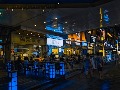 A stylish urban setting with a bustling night scene outside various cafes and restaurants. Neon and LED signs in different languages illuminate the area, casting vibrant reflections on the polished surfaces. Groups of people sit at tables conversing, while others walk along the pedestrian area, creating a lively and social atmosphere.