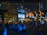 A stylish urban setting with a bustling night scene outside various cafes and restaurants. Neon and LED signs in different languages illuminate the area, casting vibrant reflections on the polished surfaces. Groups of people sit at tables conversing, while others walk along the pedestrian area, creating a lively and social atmosphere.