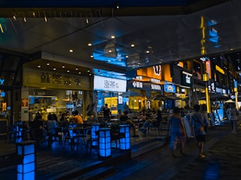 A stylish urban setting with a bustling night scene outside various cafes and restaurants. Neon and LED signs in different languages illuminate the area, casting vibrant reflections on the polished surfaces. Groups of people sit at tables conversing, while others walk along the pedestrian area, creating a lively and social atmosphere.