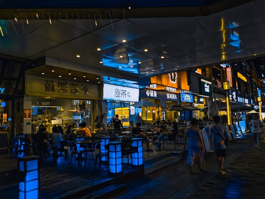 A stylish urban setting with a bustling night scene outside various cafes and restaurants. Neon and LED signs in different languages illuminate the area, casting vibrant reflections on the polished surfaces. Groups of people sit at tables conversing, while others walk along the pedestrian area, creating a lively and social atmosphere.