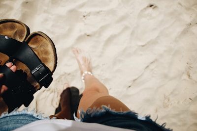 A person holding a pair of sandals while standing on sandy beach. The individual is barefoot, wearing denim shorts, and an anklet. The sand appears soft and undisturbed.