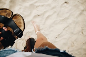 A person holding a pair of sandals while standing on sandy beach. The individual is barefoot, wearing denim shorts, and an anklet. The sand appears soft and undisturbed.