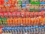 Shelves filled with various packages of instant noodles, organized by color, including red, orange, blue, and green packaging. Each shelf contains different brands and flavors, with clear branding and images of noodles on the packaging.