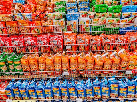 Shelves filled with various packages of instant noodles, organized by color, including red, orange, blue, and green packaging. Each shelf contains different brands and flavors, with clear branding and images of noodles on the packaging.