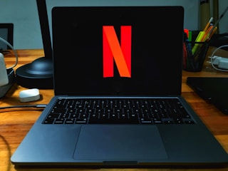 A laptop on a wooden desk with the Netflix logo prominently displayed on its screen. The area is well-organized, featuring a desk lamp, a container holding a variety of pens and pencils, and several cords. The setting suggests a workspace that combines functionality with entertainment.