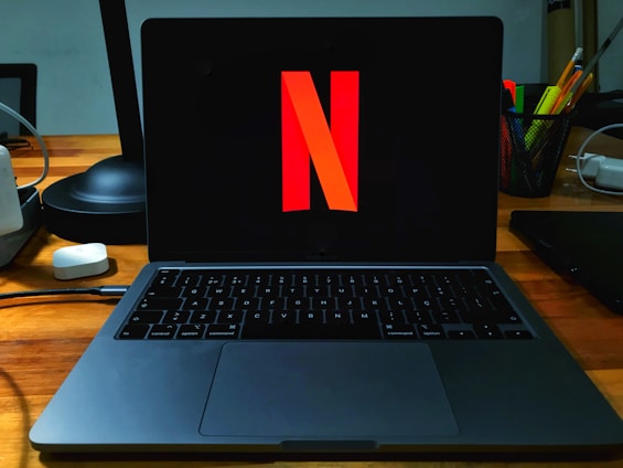 A laptop on a wooden desk with the Netflix logo prominently displayed on its screen. The area is well-organized, featuring a desk lamp, a container holding a variety of pens and pencils, and several cords. The setting suggests a workspace that combines functionality with entertainment.