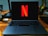 A laptop on a wooden desk with the Netflix logo prominently displayed on its screen. The area is well-organized, featuring a desk lamp, a container holding a variety of pens and pencils, and several cords. The setting suggests a workspace that combines functionality with entertainment.