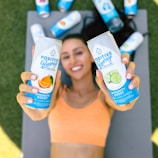 A person is holding two cans of 'Positive Beverage' labeled with flavors 'Mandarin Orange' and 'Crisp Cucumber.' The individual is lying on a yoga mat outdoors, wearing an orange sports top. Other cans are scattered in the background on the grass.