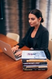 A person is working on a laptop at a wooden table. Next to them is a stack of books relating to business and success, with titles visible such as 'The Warren Buffett Way' and 'The Deals That Made The World'. The setting appears to be professional, possibly an office or study.