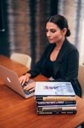 A person is working on a laptop at a wooden table. Next to them is a stack of books relating to business and success, with titles visible such as 'The Warren Buffett Way' and 'The Deals That Made The World'. The setting appears to be professional, possibly an office or study.