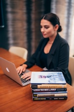 A person is working on a laptop at a wooden table. Next to them is a stack of books relating to business and success, with titles visible such as 'The Warren Buffett Way' and 'The Deals That Made The World'. The setting appears to be professional, possibly an office or study.