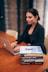 A person is working on a laptop at a wooden table. Next to them is a stack of books relating to business and success, with titles visible such as 'The Warren Buffett Way' and 'The Deals That Made The World'. The setting appears to be professional, possibly an office or study.