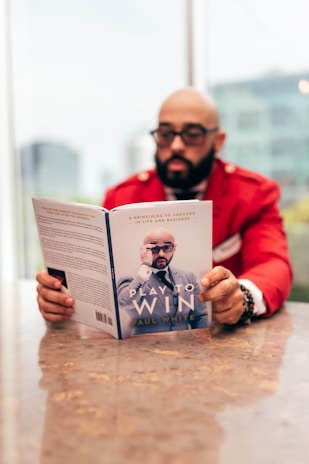 A person wearing glasses and a red jacket is sitting at a table, reading a book titled 'Play to Win' by Paul White. The background is blurred, showing a hint of a cityscape through large windows.