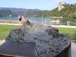 A bronze relief map focusing on the foreground, with detailed topographic features depicted, overlooking a scenic lake. In the background, a couple sits on a bench by the water, enjoying the view. Two individuals walk along a path parallel to the lake. A castle is perched on a hill surrounded by lush greenery on the right, with mountains in the distance.