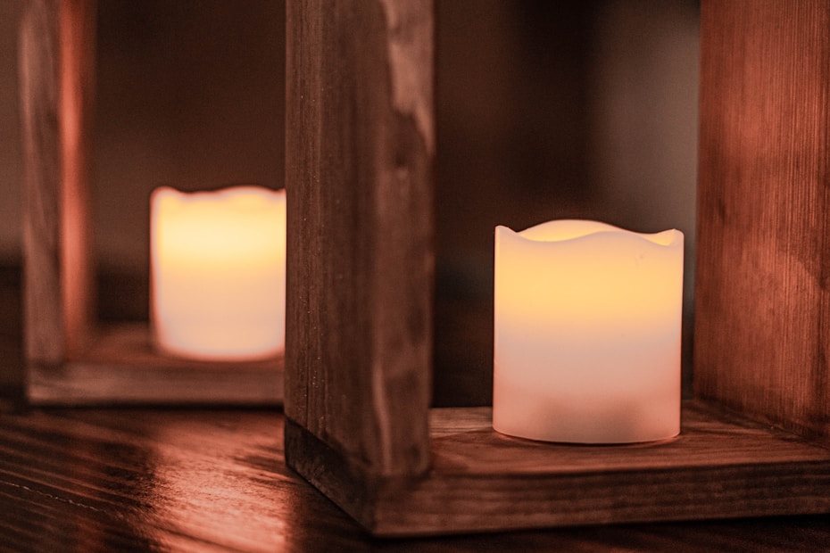 Two candles emitting a warm, soft glow are placed inside rustic, wooden lanterns. The candles are set on a wooden surface, creating a cozy and serene ambiance.