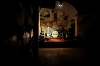 A dimly lit stage featuring a drum set with a white bass drum displaying 'The Beatles,' surrounded by several guitars. The background consists of a colorful, patchwork-style wall, creating a warm and nostalgic atmosphere. The spotlight focuses on the instruments, evoking a sense of anticipation and history.