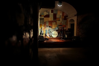 A dimly lit stage featuring a drum set with a white bass drum displaying 'The Beatles,' surrounded by several guitars. The background consists of a colorful, patchwork-style wall, creating a warm and nostalgic atmosphere. The spotlight focuses on the instruments, evoking a sense of anticipation and history.