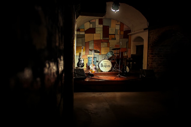 A dimly lit stage featuring a drum set with a white bass drum displaying 'The Beatles,' surrounded by several guitars. The background consists of a colorful, patchwork-style wall, creating a warm and nostalgic atmosphere. The spotlight focuses on the instruments, evoking a sense of anticipation and history.
