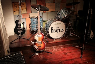 A vintage band setup with musical instruments including two electric guitars and a drum set prominently featuring 'The Beatles' logo. The scene suggests a tribute or homage to the iconic band, with various signatures and notes displayed on the wall behind the instruments. The wooden stage and warm lighting create a nostalgic atmosphere.