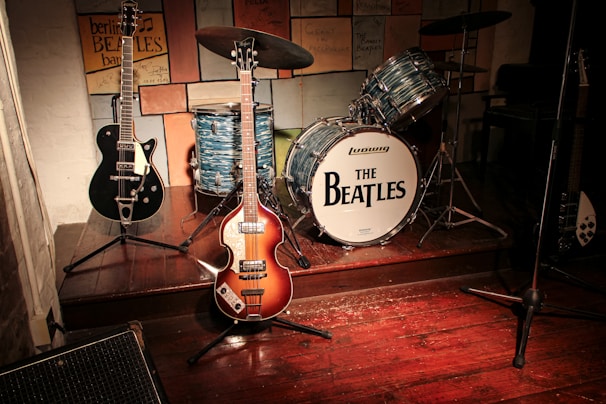A vintage band setup with musical instruments including two electric guitars and a drum set prominently featuring 'The Beatles' logo. The scene suggests a tribute or homage to the iconic band, with various signatures and notes displayed on the wall behind the instruments. The wooden stage and warm lighting create a nostalgic atmosphere.