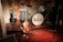 A vintage band setup with musical instruments including two electric guitars and a drum set prominently featuring 'The Beatles' logo. The scene suggests a tribute or homage to the iconic band, with various signatures and notes displayed on the wall behind the instruments. The wooden stage and warm lighting create a nostalgic atmosphere.