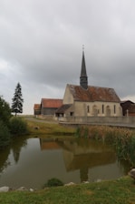 A quaint rural church with a tall steeple is surrounded by a serene landscape. The church is reflected in a small pond, adding to the peaceful ambiance. The building has a weathered, rustic appearance with patches of moss on the roof. Nearby are a few trees and a small path leading to the church.