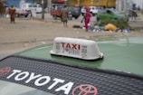 A street scene featuring a Toyota taxi with the word 'TAXI' prominently displayed on a sign on its roof. In the background, several people walk near parked cars amidst scattered garbage and debris. The environment appears urban, with buildings and vehicles visible in the distance.