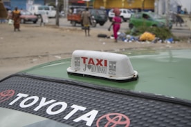 A street scene featuring a Toyota taxi with the word 'TAXI' prominently displayed on a sign on its roof. In the background, several people walk near parked cars amidst scattered garbage and debris. The environment appears urban, with buildings and vehicles visible in the distance.
