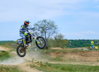 A motocross rider wearing protective gear and a helmet is caught mid-air as their bike leaps over a dirt track. The scene takes place in an open field with grassy areas and a few small trees. In the background, a person in a high-visibility vest stands near the track, holding a tool, possibly maintaining or coordinating the event.