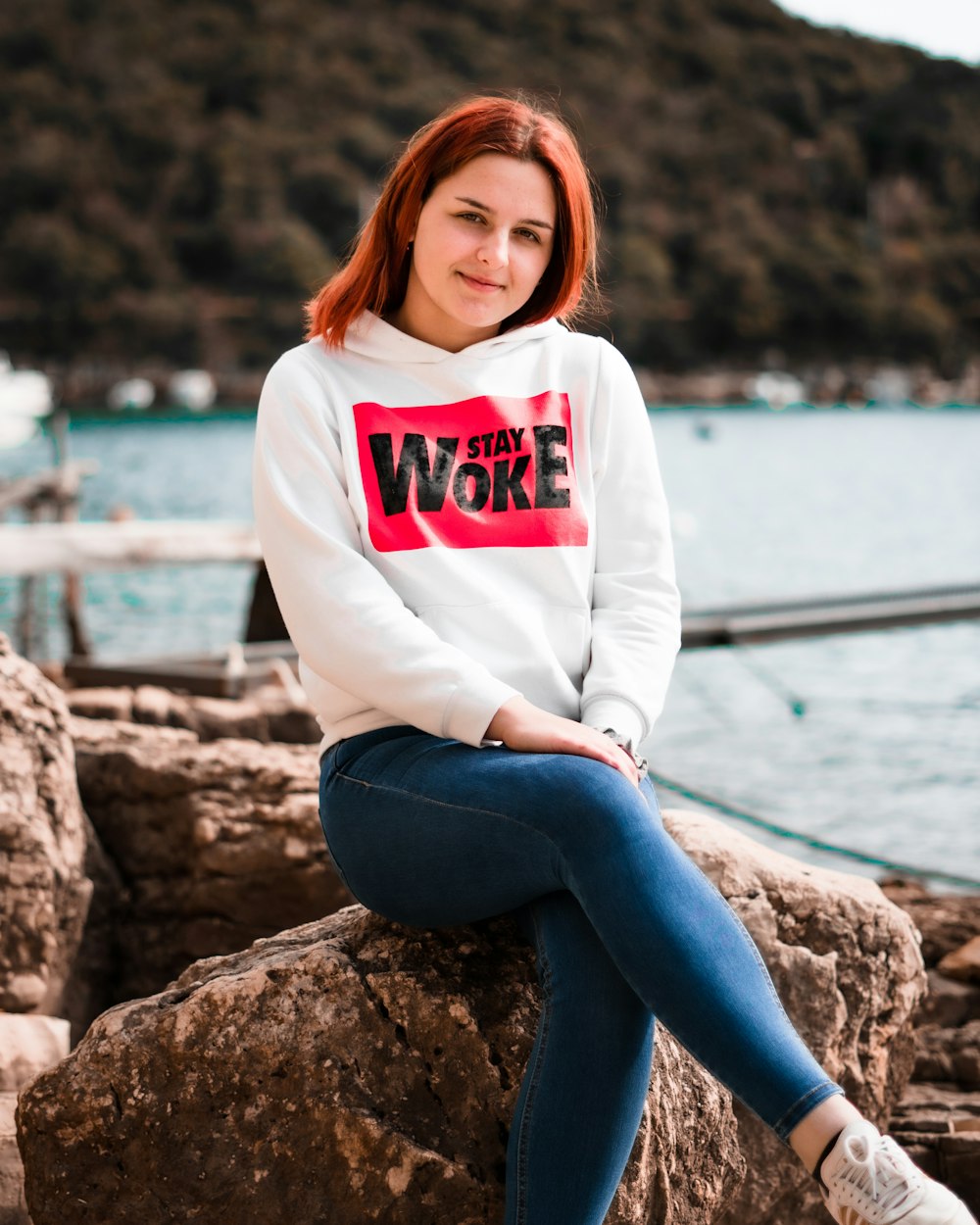 woman in white hoodie and blue denim jeans sitting on rock near body of water during